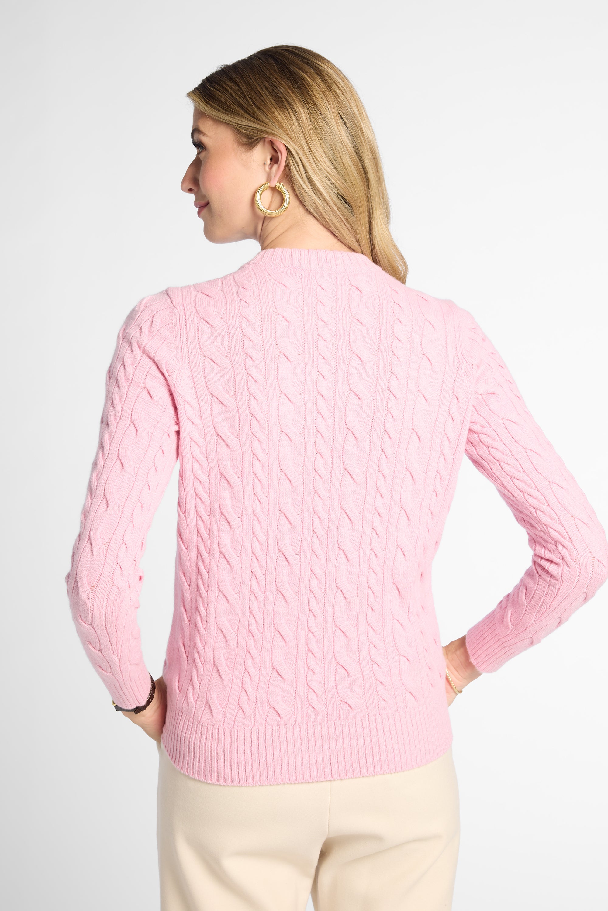 A woman shown from the back wearing a light pink cable-knit sweater and cream pants against a plain light gray studio background.