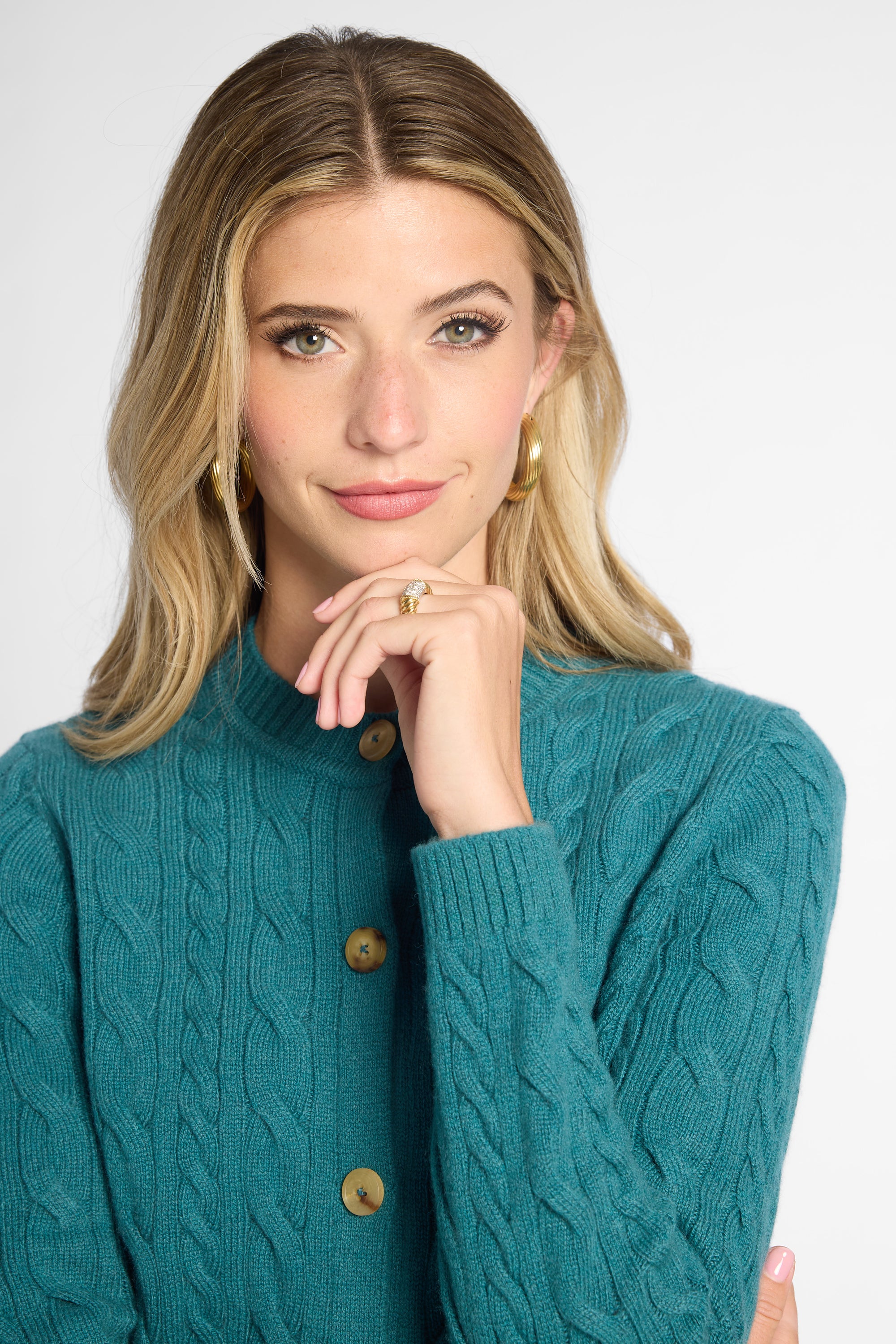 A woman with blonde hair wearing a teal cable-knit button-up cardigan sweater, styled with gold hoop earrings and a gold ring, against a plain light gray background.