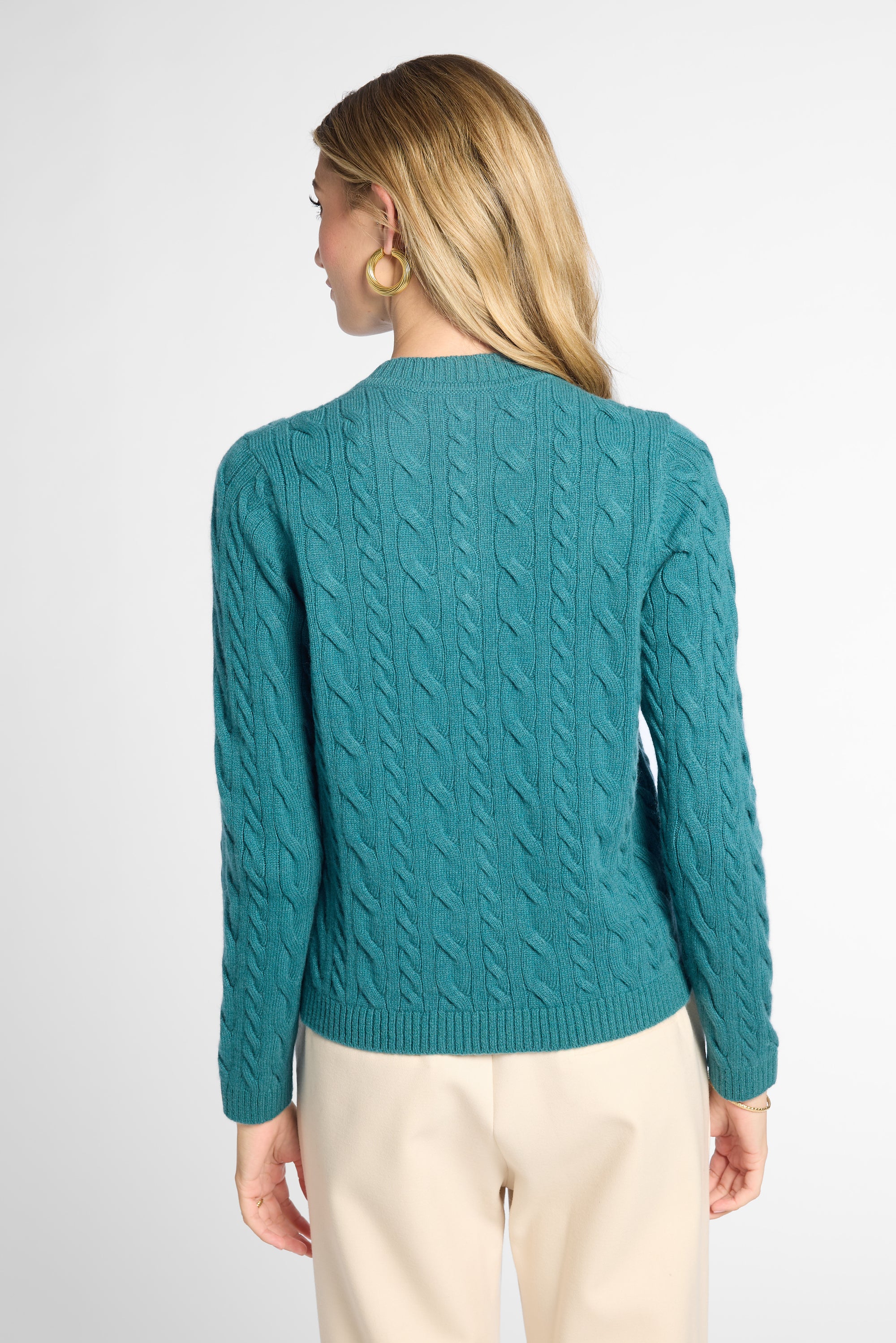 A woman shown from the back wearing a teal cable-knit sweater and cream pants against a light gray studio background.