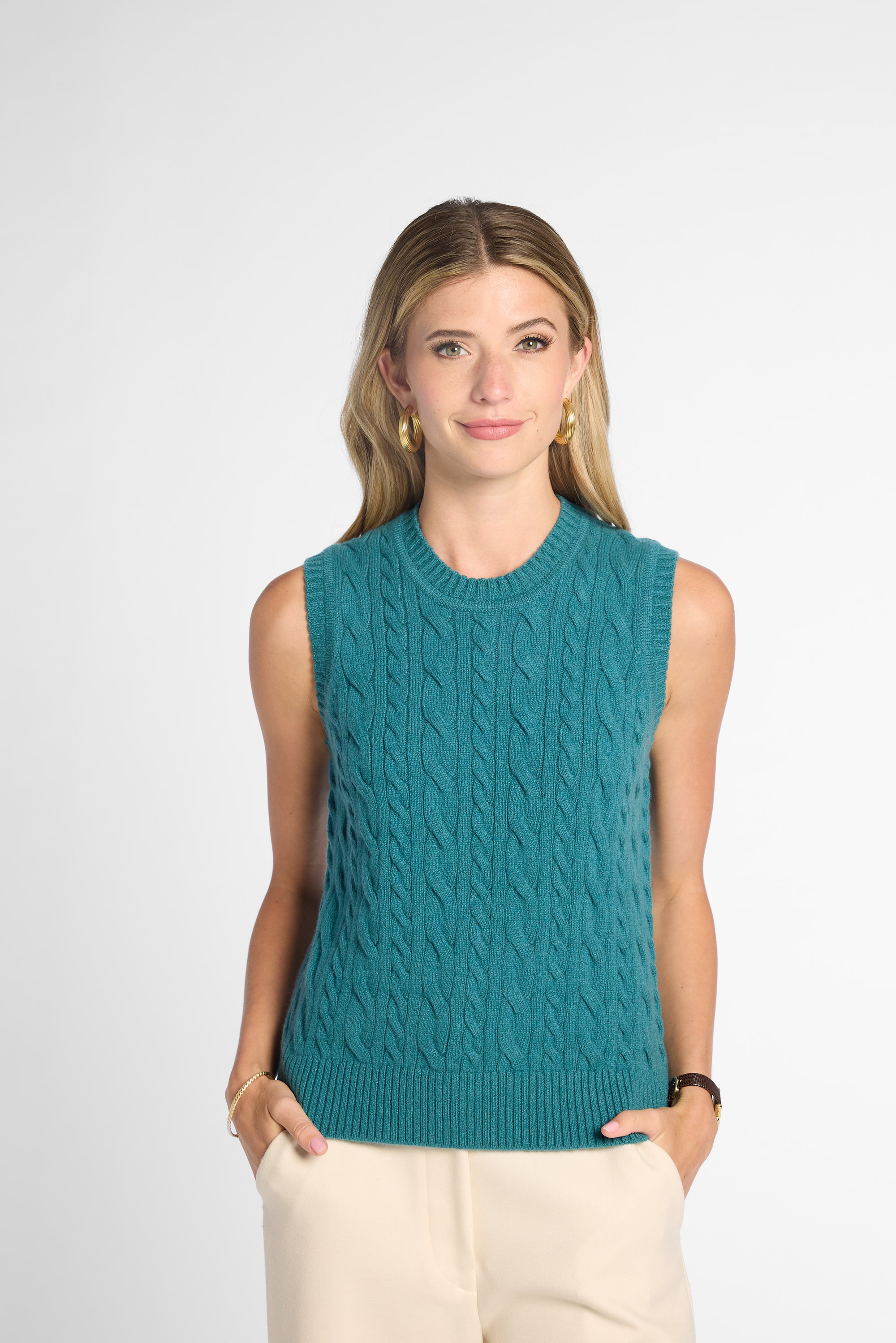 A woman modeling a sleeveless teal cable-knit sweater vest with cream pants, standing against a light gray studio background.