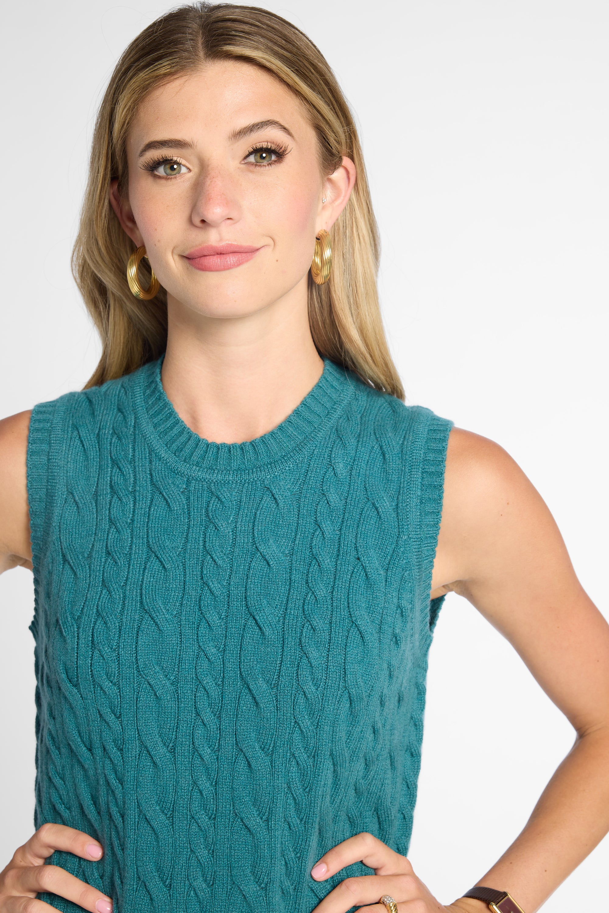 A woman wearing a sleeveless cable-knit sweater vest in teal, paired with gold hoop earrings, against a plain light gray studio background.