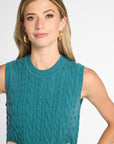 A woman wearing a sleeveless cable-knit sweater vest in teal, paired with gold hoop earrings, against a plain light gray studio background.