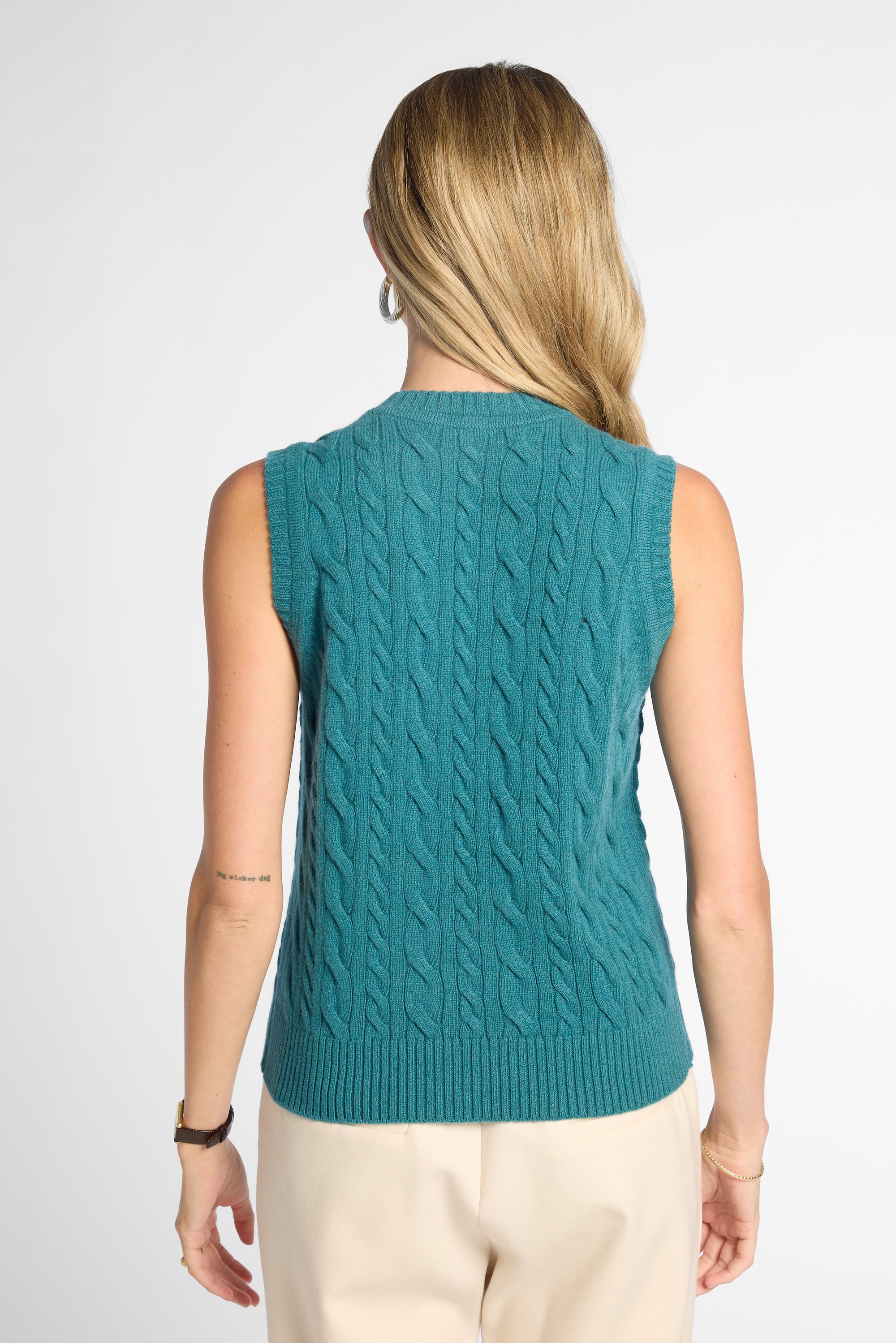 A woman shown from the back wearing a sleeveless teal cable-knit sweater vest over a light beige top and pants, against a plain light gray studio background.