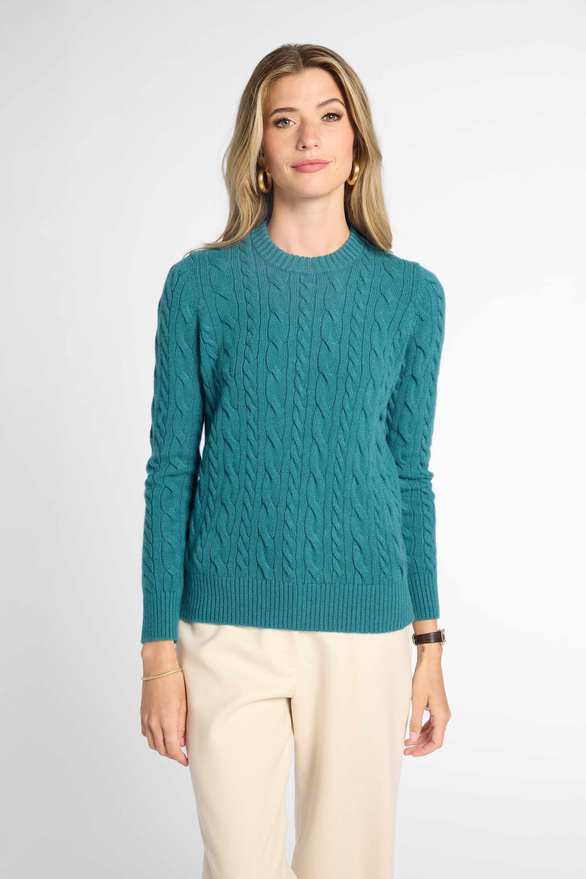 A woman modeling a teal cable-knit crewneck sweater with cream trousers, standing against a light gray studio background.