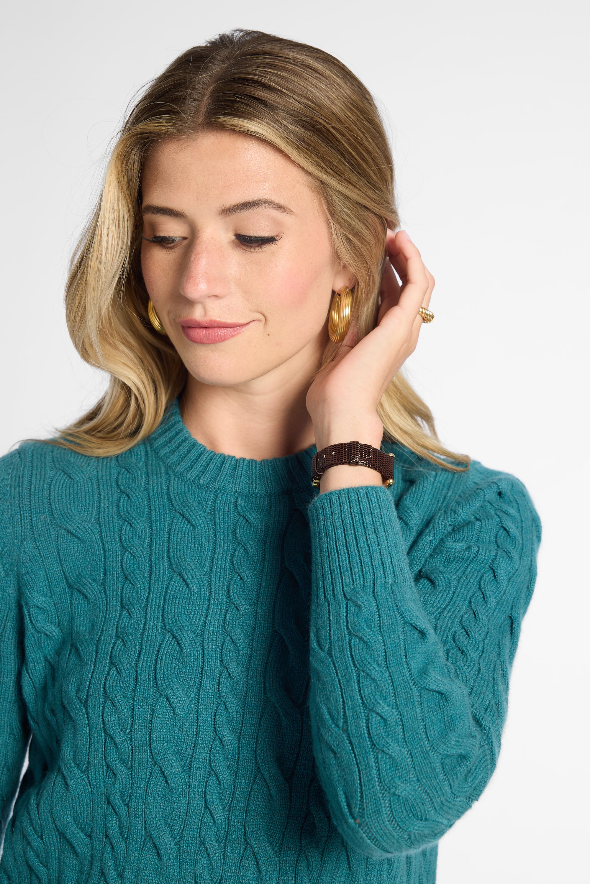 A woman wearing a teal cable-knit sweater, gold hoop earrings, and a brown leather watch, posing against a light gray studio background.
