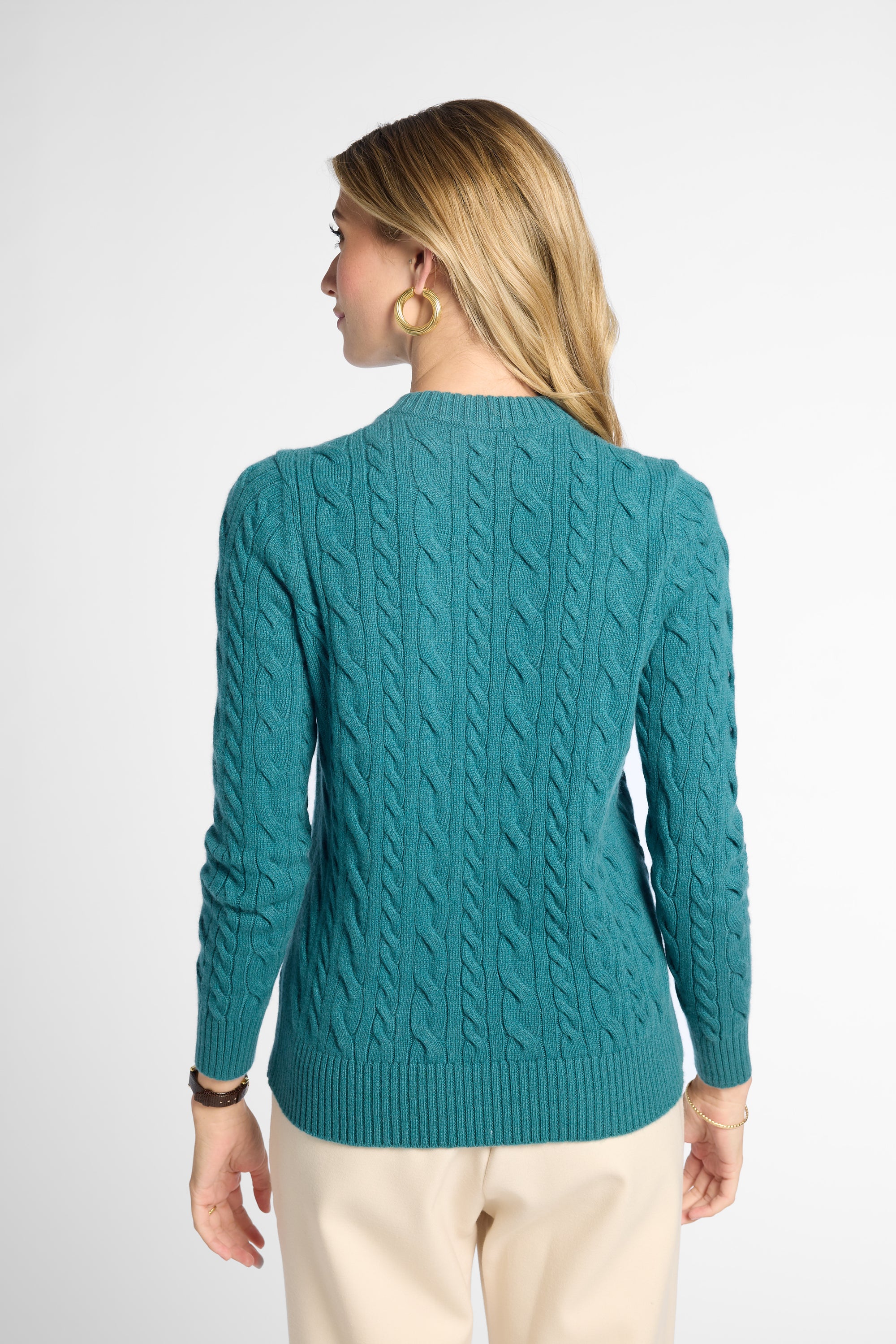 A woman shown from the back wearing a teal cable-knit sweater and cream pants against a light gray studio background.