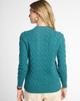 A woman shown from the back wearing a teal cable-knit sweater and cream pants against a light gray studio background.