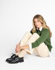 A woman sits on the floor against a white studio background, wearing a green knit sweater over a white collared shirt, cream trousers, beige socks, and black lace-up shoes.