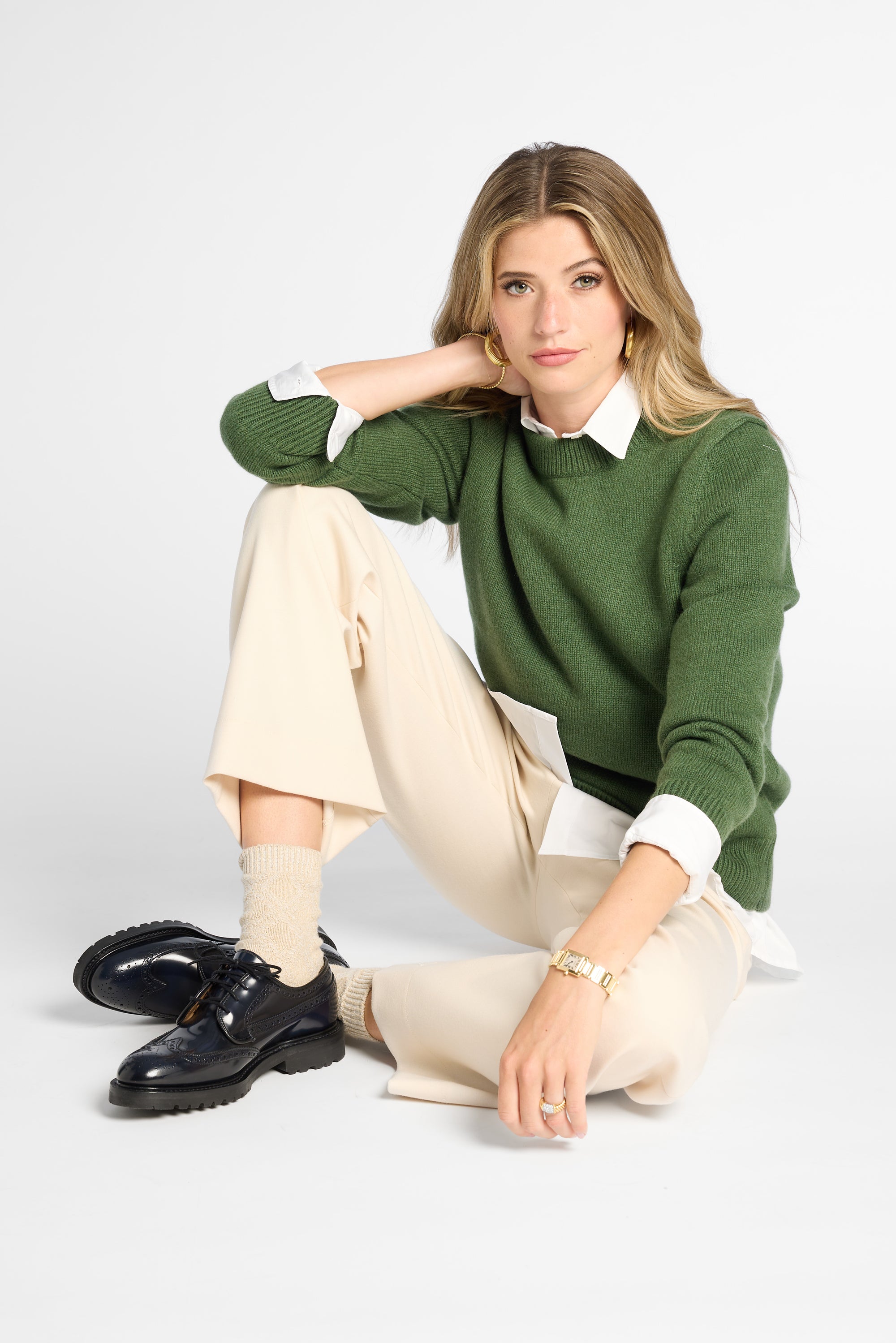 A woman sits cross-legged on a light gray studio floor wearing a green knit sweater over a white collared shirt, cream trousers, beige socks, and navy shoes.
