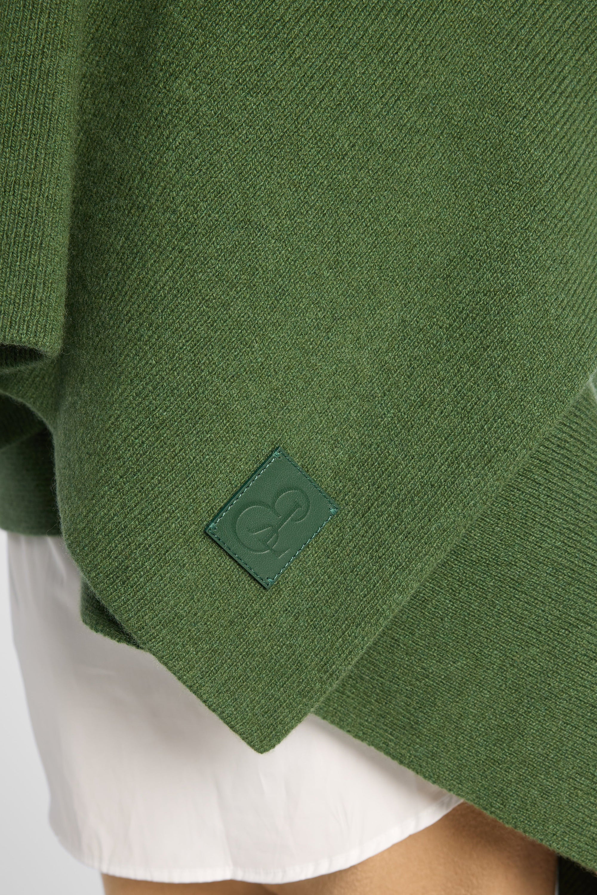 Close view of women wearing a Forest Green cashmere wrap over a white shirt and tan pants in front of a white background. Embossed green leather patch shown on the corner.