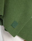 Close view of women wearing a Forest Green cashmere wrap over a white shirt and tan pants in front of a white background. Embossed green leather patch shown on the corner.