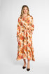 A woman modeling a long-sleeve midi dress with a floral print in orange, pink, and brown on a cream background, paired with dark brown pointed-toe flats, against a light gray studio background.