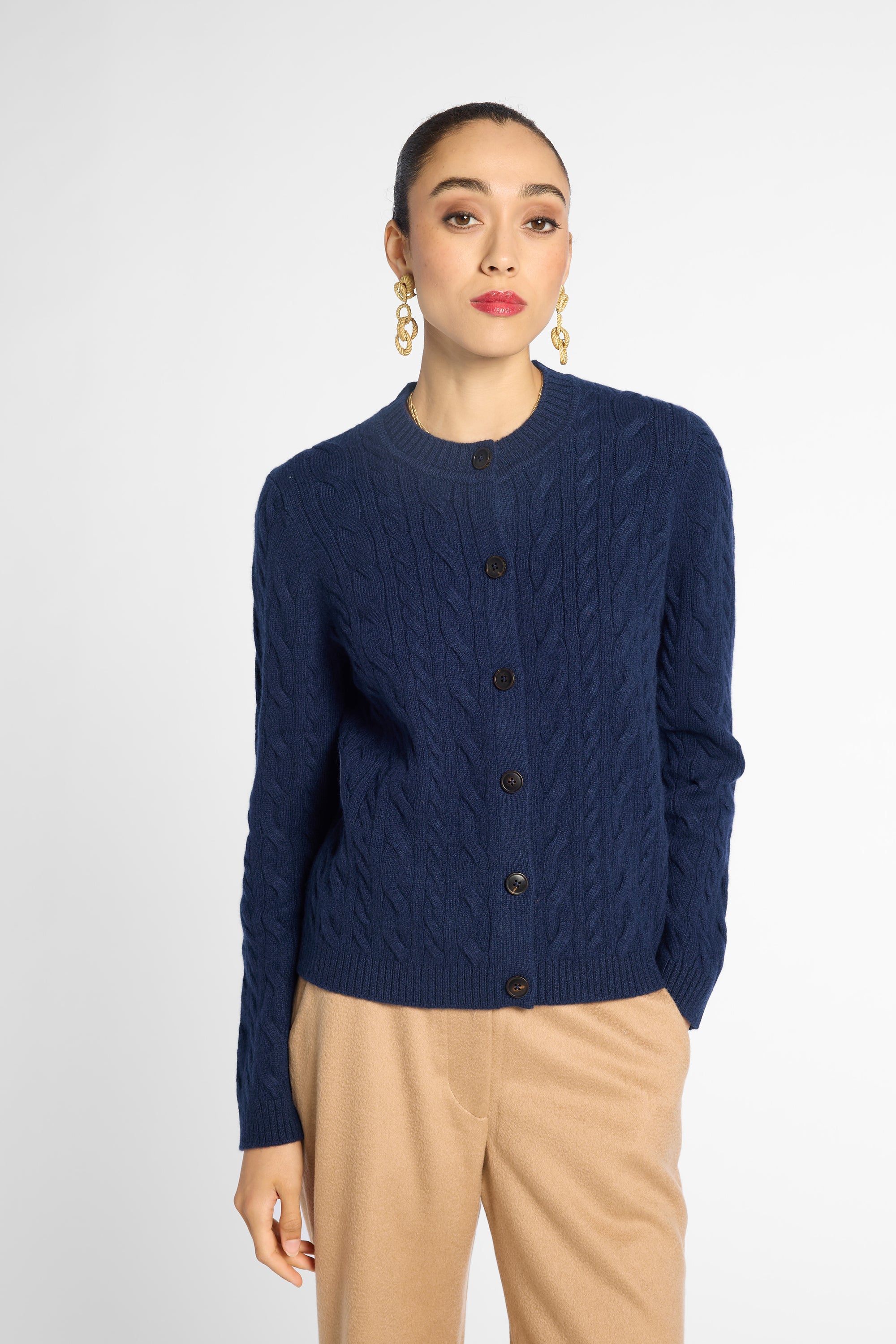 A woman modeling a navy blue cable-knit cardigan sweater with dark buttons, paired with beige trousers, against a light gray studio background.