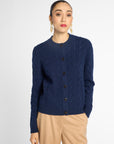 A woman modeling a navy blue cable-knit cardigan sweater with dark buttons, paired with beige trousers, against a light gray studio background.
