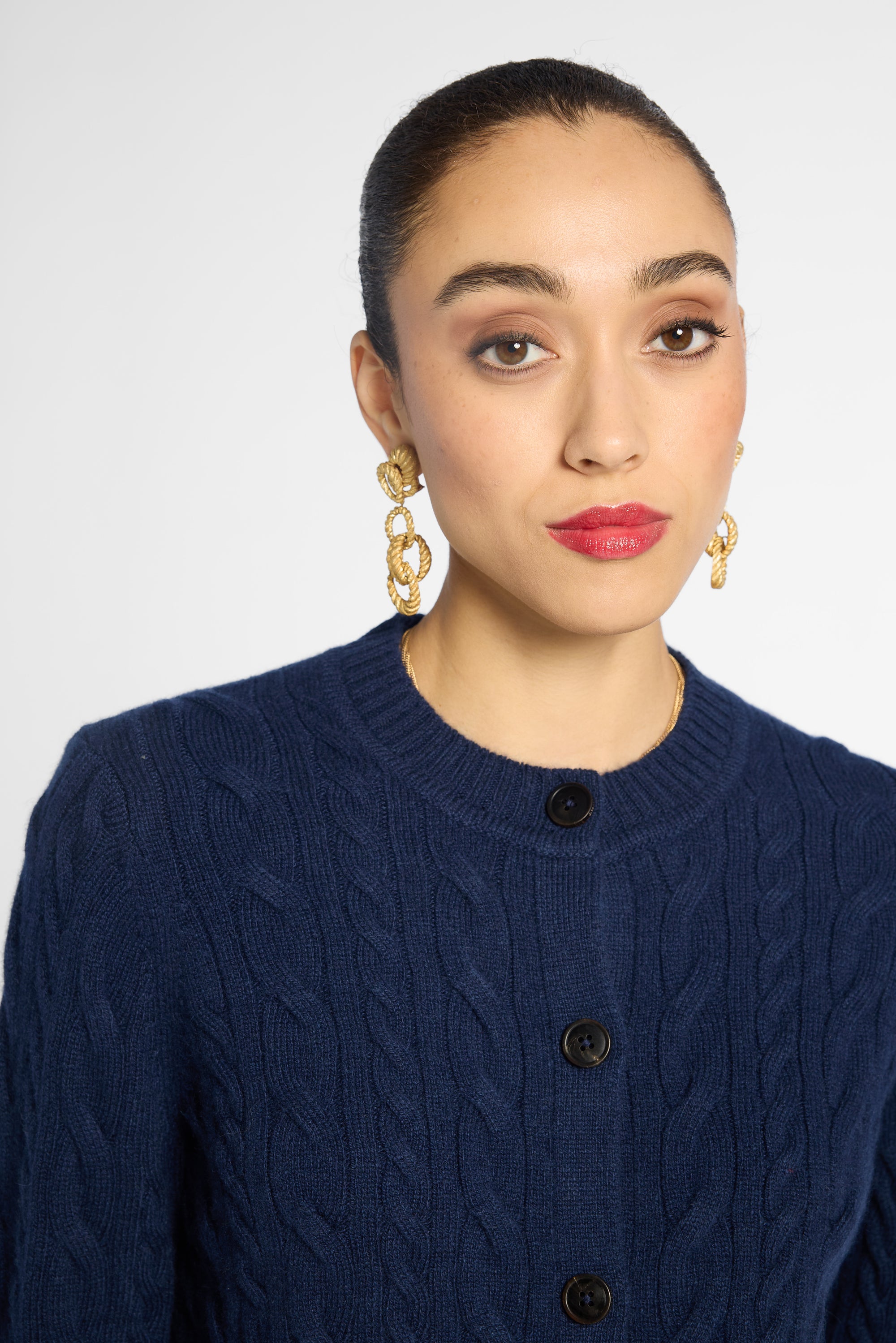 A close-up of a woman wearing a navy blue cable-knit cardigan sweater and gold chain-link drop earrings against a light gray background.