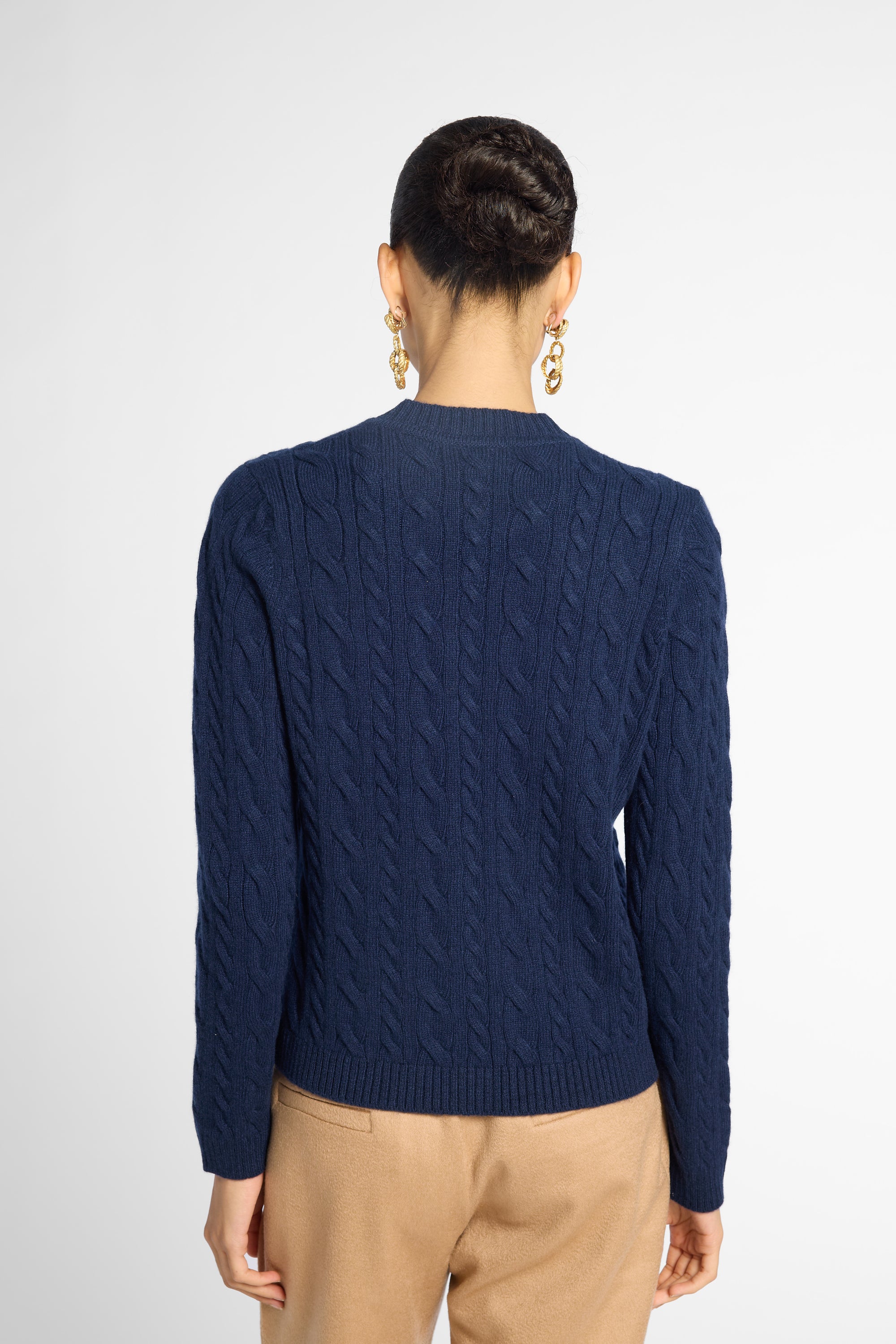 A woman viewed from the back wearing a navy blue cable-knit sweater and tan trousers, against a light gray studio background.