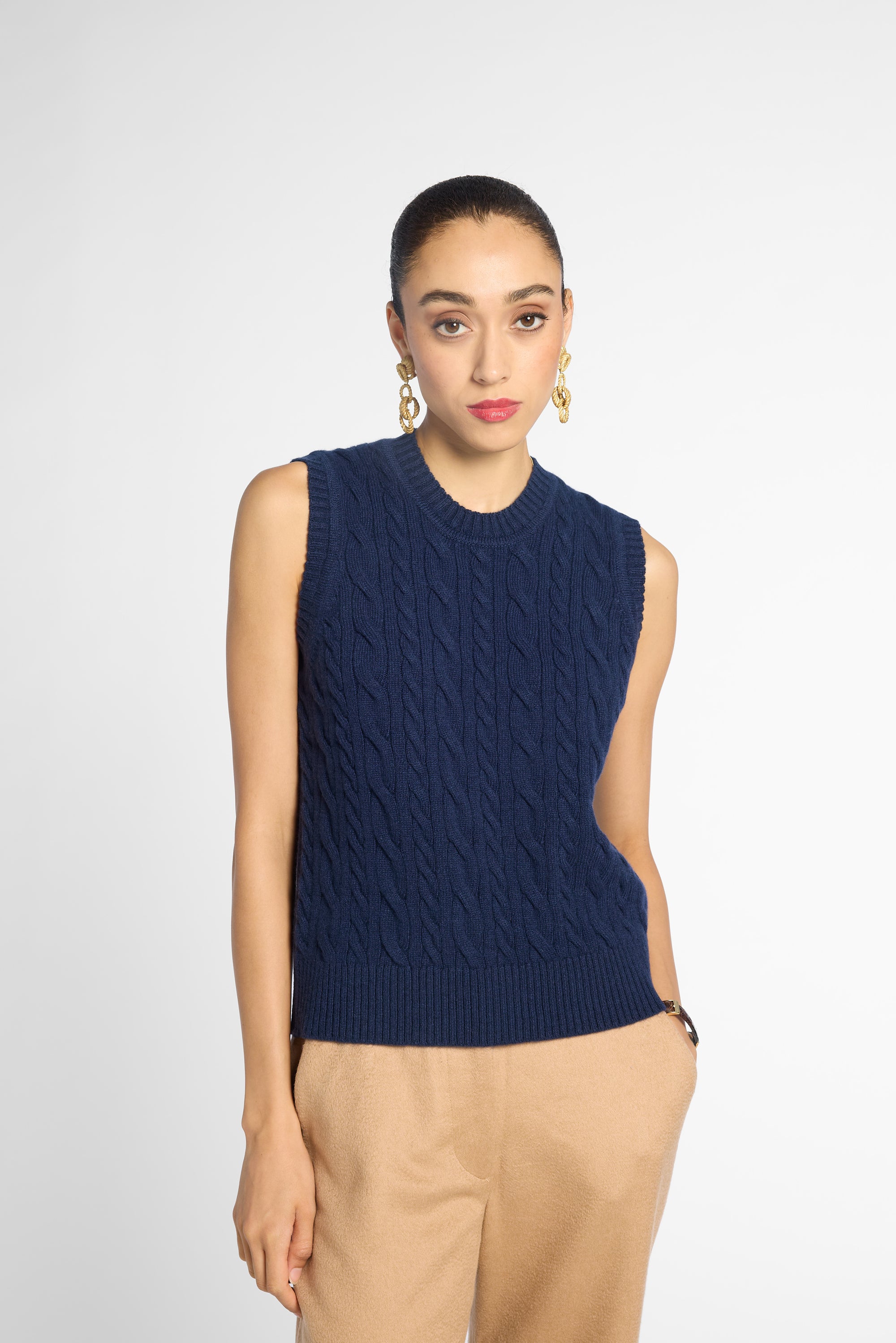 A woman modeling a sleeveless navy blue cable-knit sweater vest paired with beige trousers, shot against a light gray studio background.