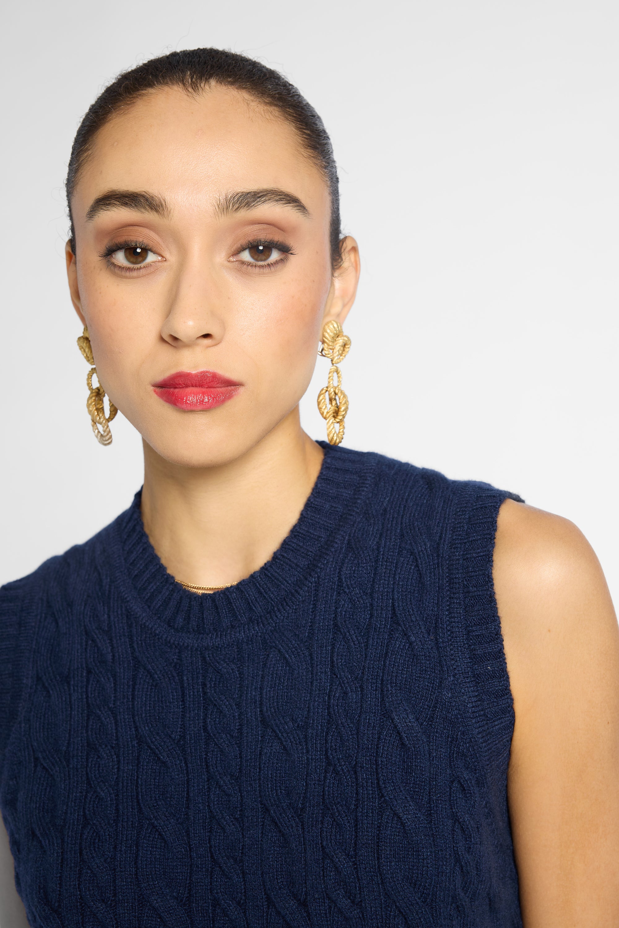 A close-up portrait of a model wearing chunky gold chain-link drop earrings and a navy blue cable-knit sleeveless sweater vest against a light gray studio background.