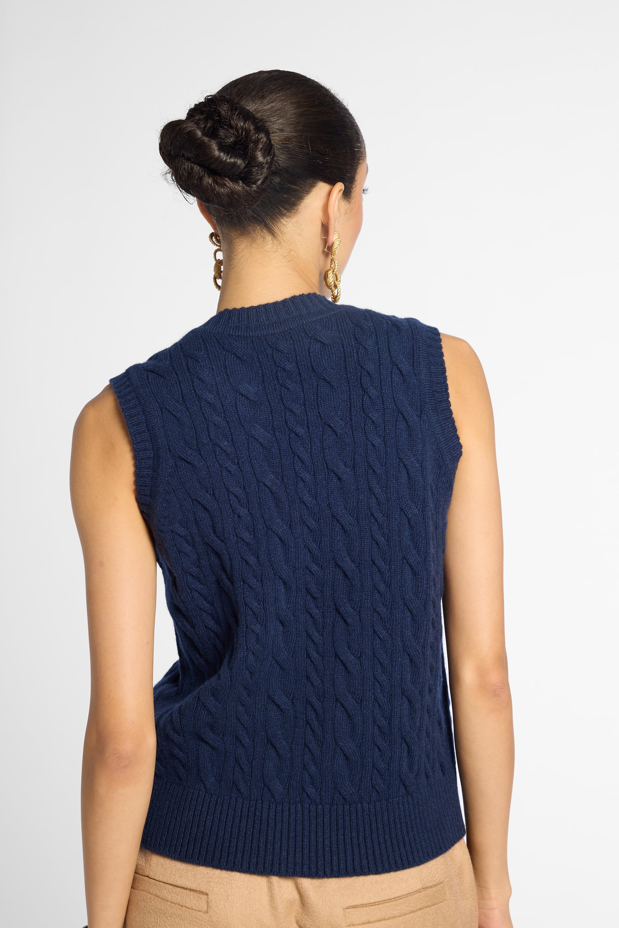 A woman shown from the back wearing a sleeveless navy blue cable-knit sweater vest over a beige top, against a plain light gray studio background.