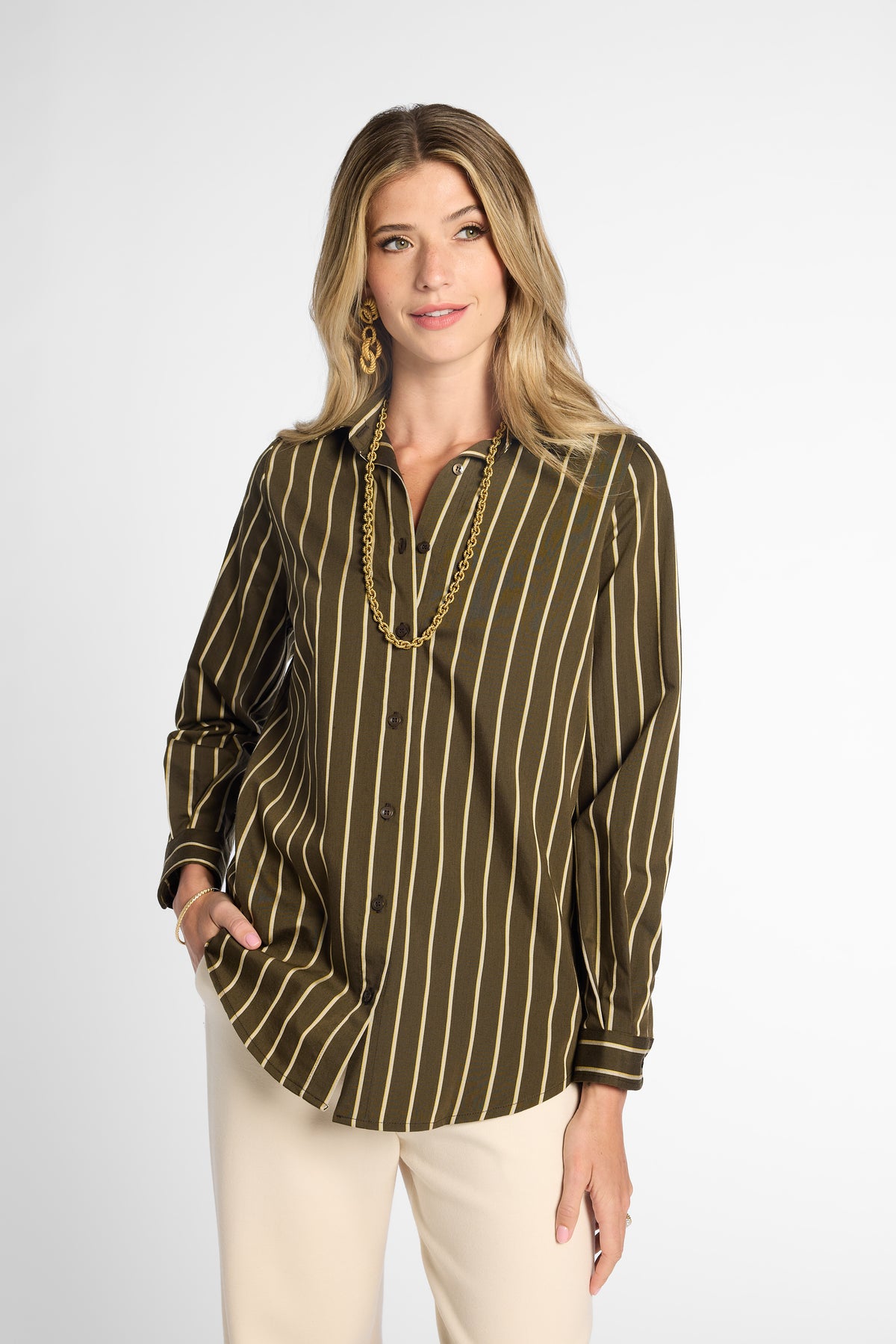 A woman modeling a long-sleeve button-up blouse with vertical olive green and tan stripes, paired with cream pants, against a light gray studio background.
