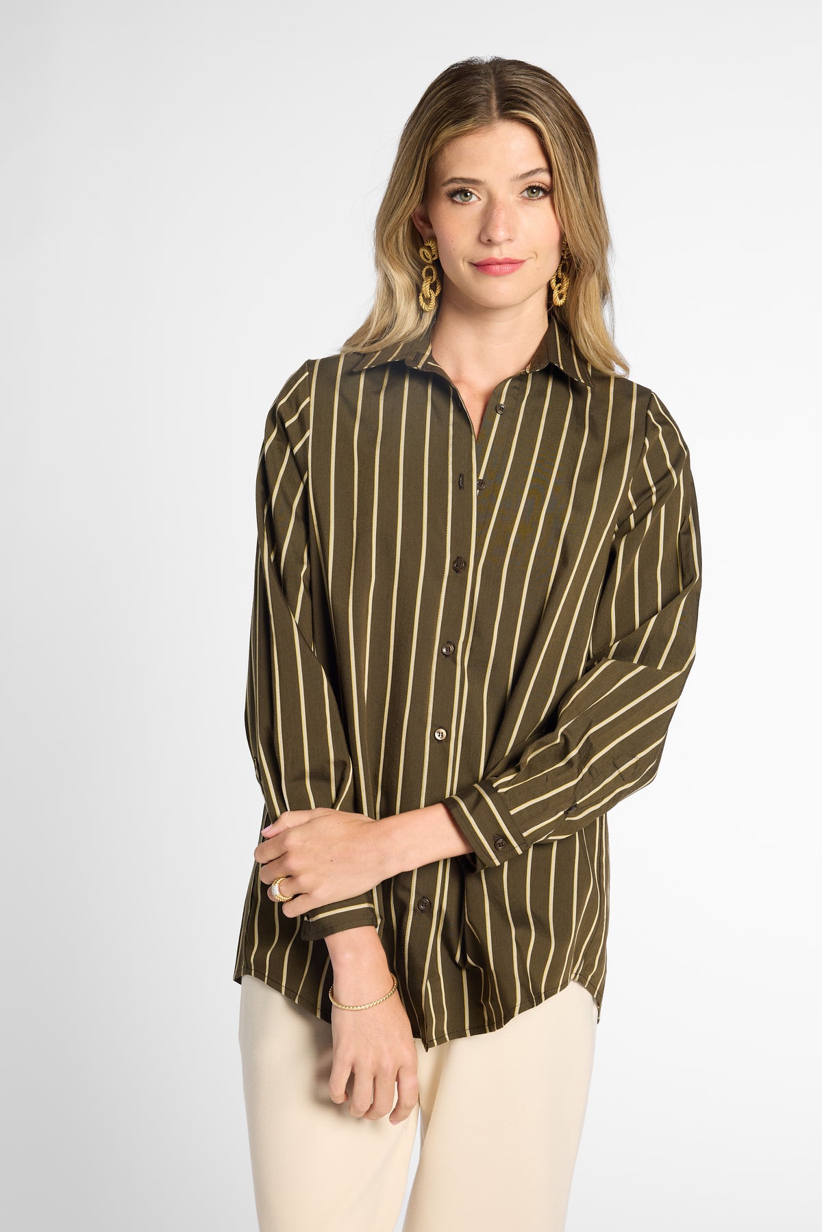 A woman modeling a long-sleeve button-up shirt with vertical olive green and beige stripes, paired with cream pants, against a light gray studio background.
