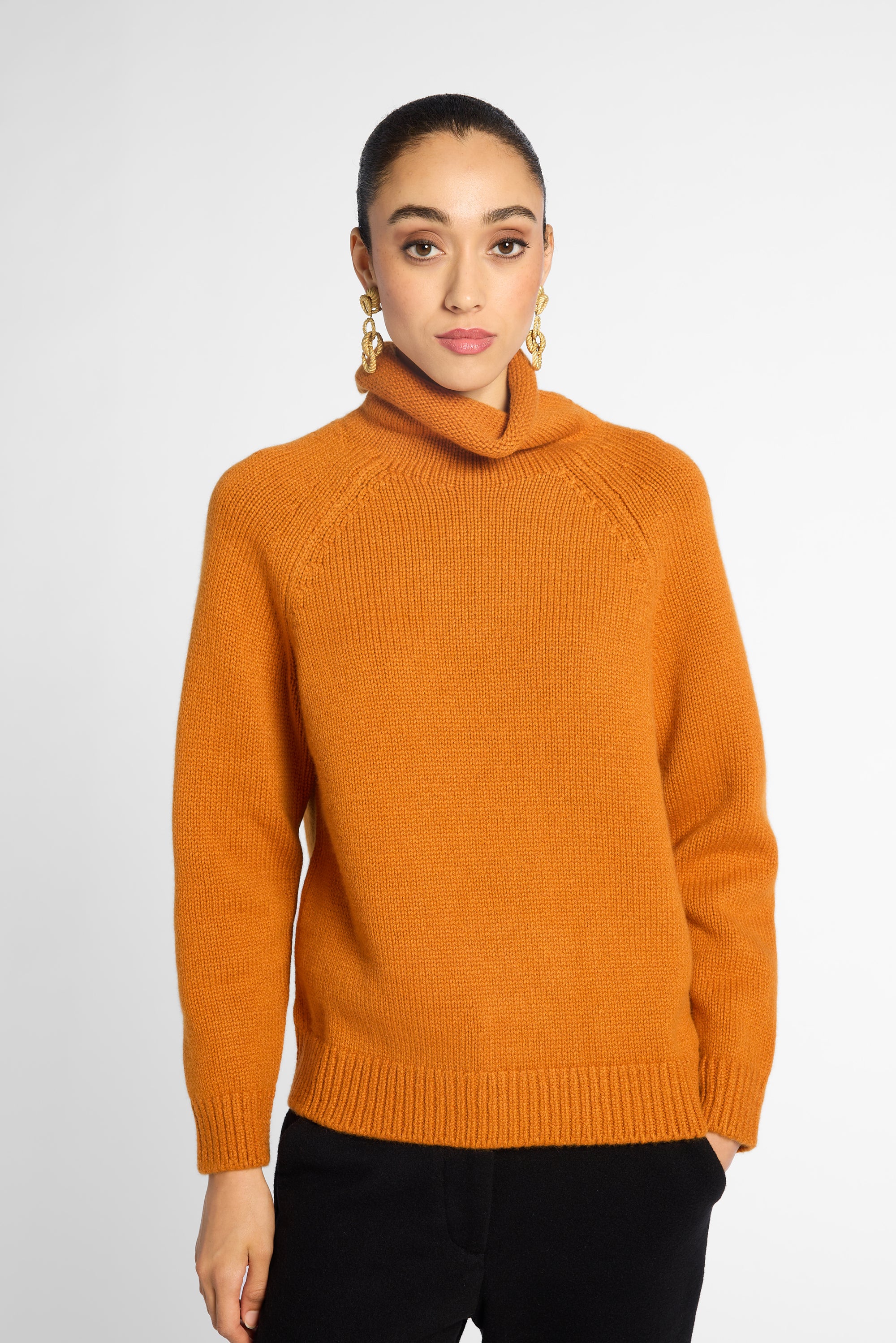 A woman wearing a bright orange knit cowl-neck sweater with raglan sleeves, paired with black pants, against a light gray studio background.