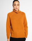 A woman wearing a bright orange knit cowl-neck sweater with raglan sleeves, paired with black pants, against a light gray studio background.