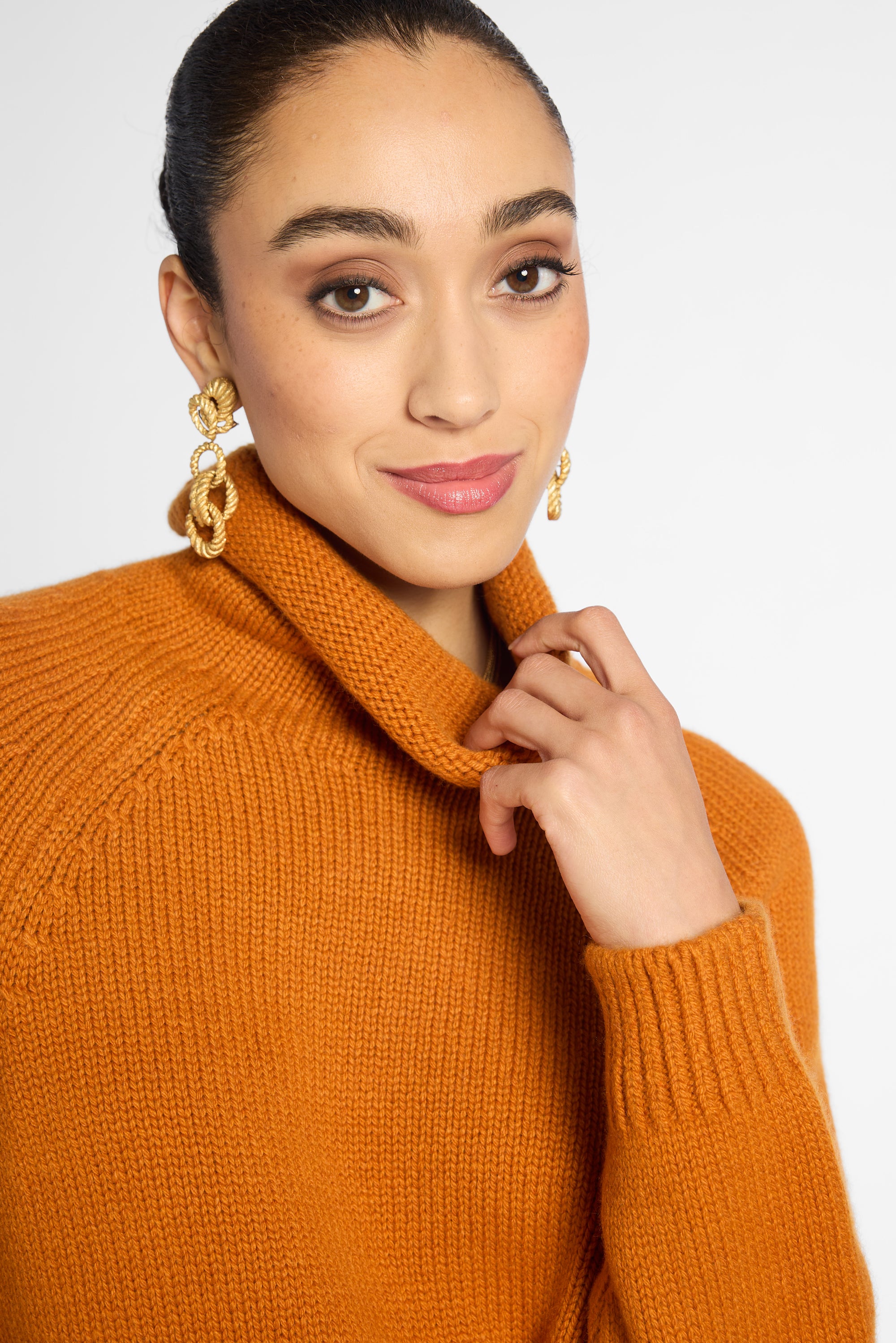 A close-up of a woman wearing a chunky knit turtleneck sweater in a warm camel orange color, paired with chunky gold chain drop earrings, against a plain light gray background.
