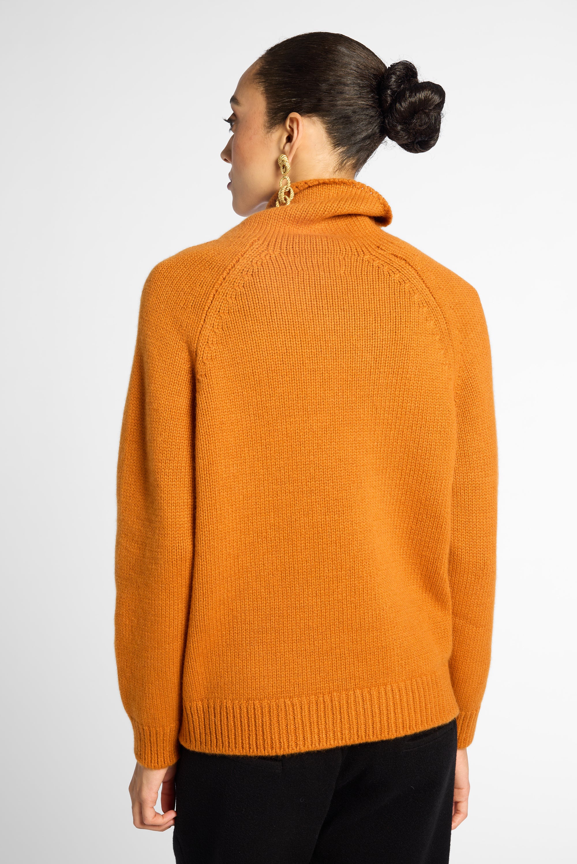 A woman shown from the back wearing a bright orange knit sweater with a turtleneck and raglan sleeves, paired with black pants, against a light gray studio background.