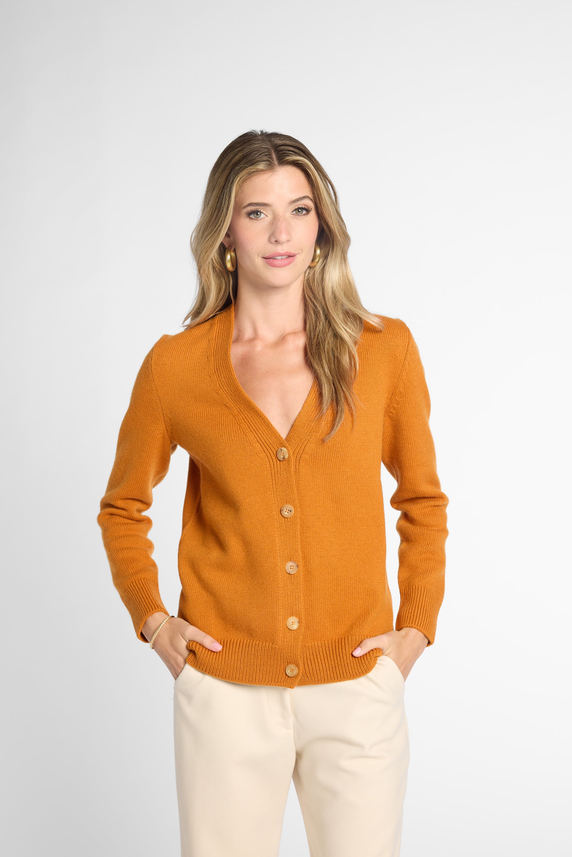 A woman modeling an orange knit cardigan sweater with a V-neck and button-front closure, paired with cream pants, against a light gray studio background.
