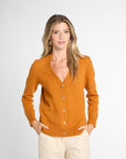 A woman modeling an orange knit cardigan sweater with a V-neck and button-front closure, paired with cream pants, against a light gray studio background.