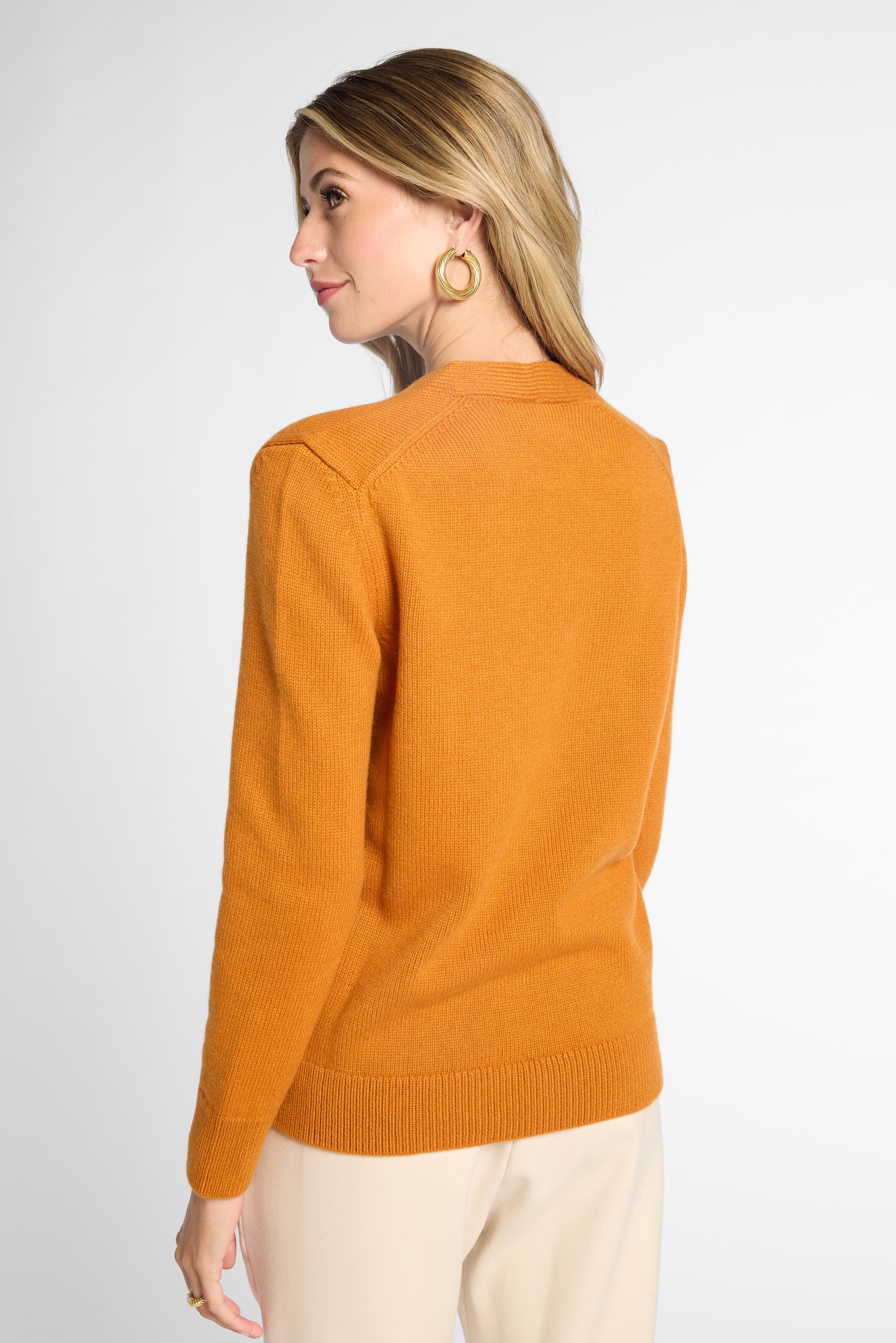A woman shown from the back wearing a solid orange knit sweater and cream-colored pants, photographed against a clean white studio background.