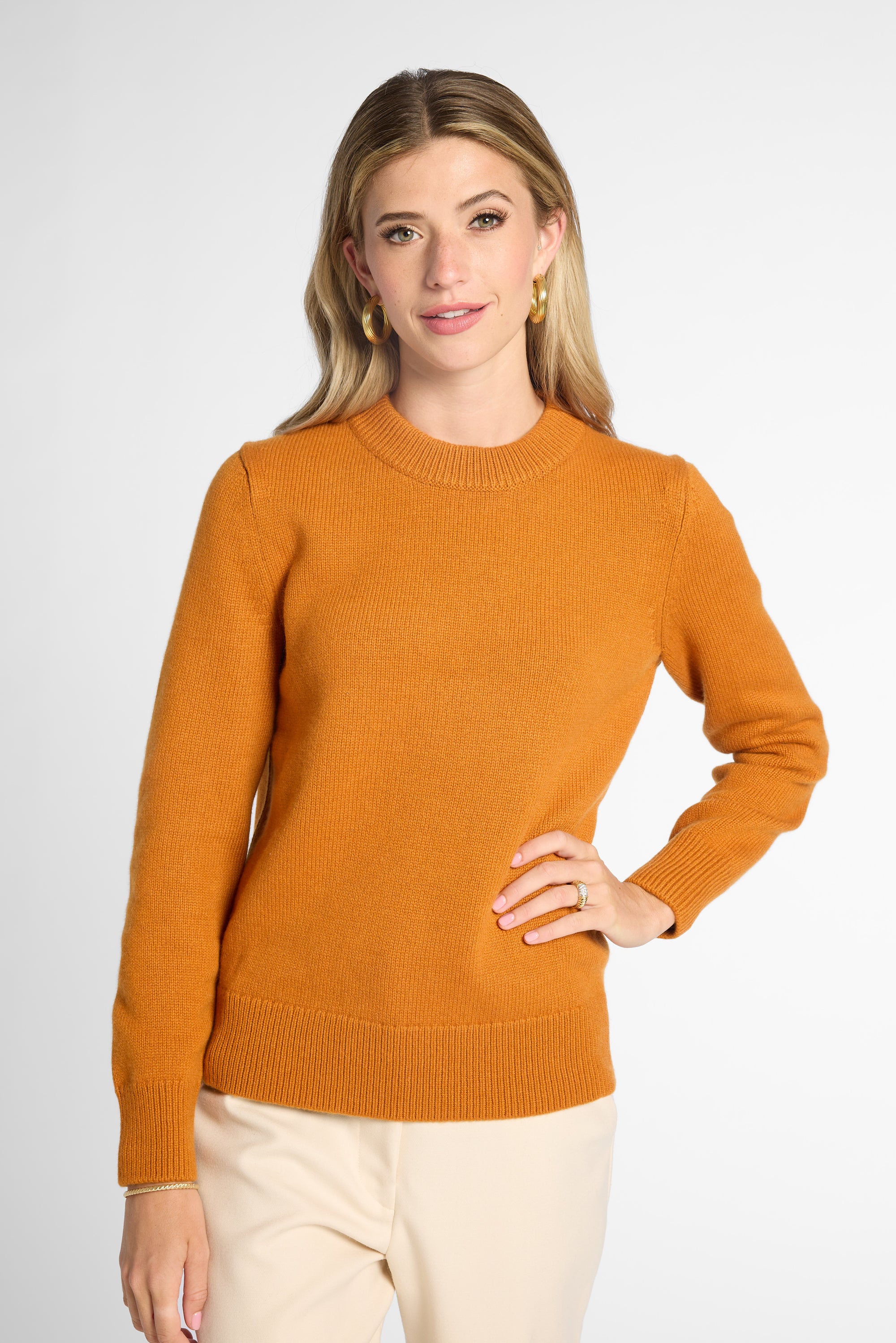 A woman wearing an orange knit crewneck sweater, gold hoop earrings, and a delicate bracelet, photographed against a clean light gray studio background.