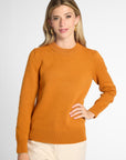 A woman wearing an orange knit crewneck sweater, gold hoop earrings, and a delicate bracelet, photographed against a clean light gray studio background.