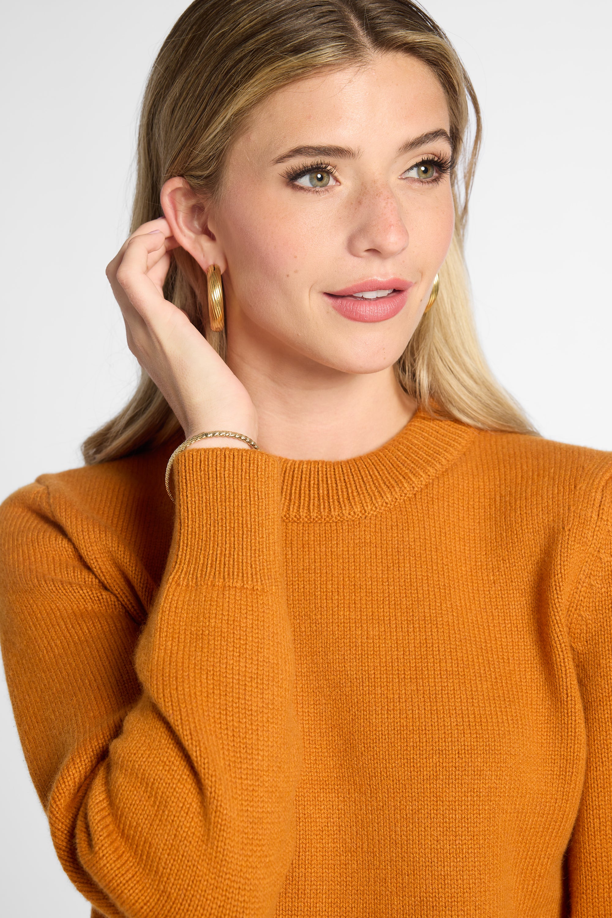 Closeup of a woman wearing an orange knit crewneck sweater, gold hoop earrings, and a delicate bracelet, photographed against a clean light gray studio background.