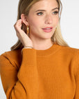 Closeup of a woman wearing an orange knit crewneck sweater, gold hoop earrings, and a delicate bracelet, photographed against a clean light gray studio background.