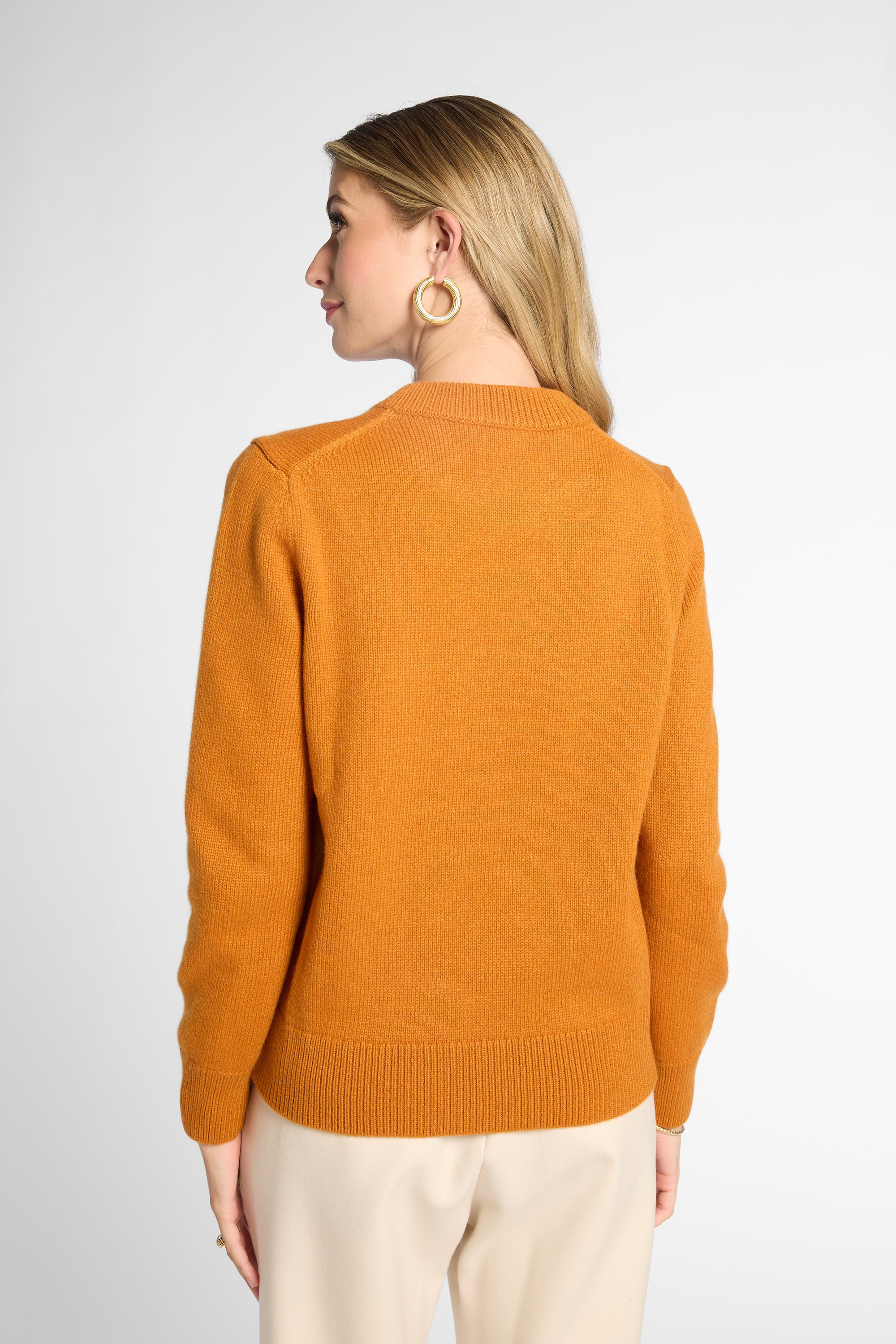 A woman shown from the back wearing an orang knit sweater and cream trousers against a light gray studio background.