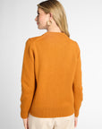 A woman shown from the back wearing an orang knit sweater and cream trousers against a light gray studio background.