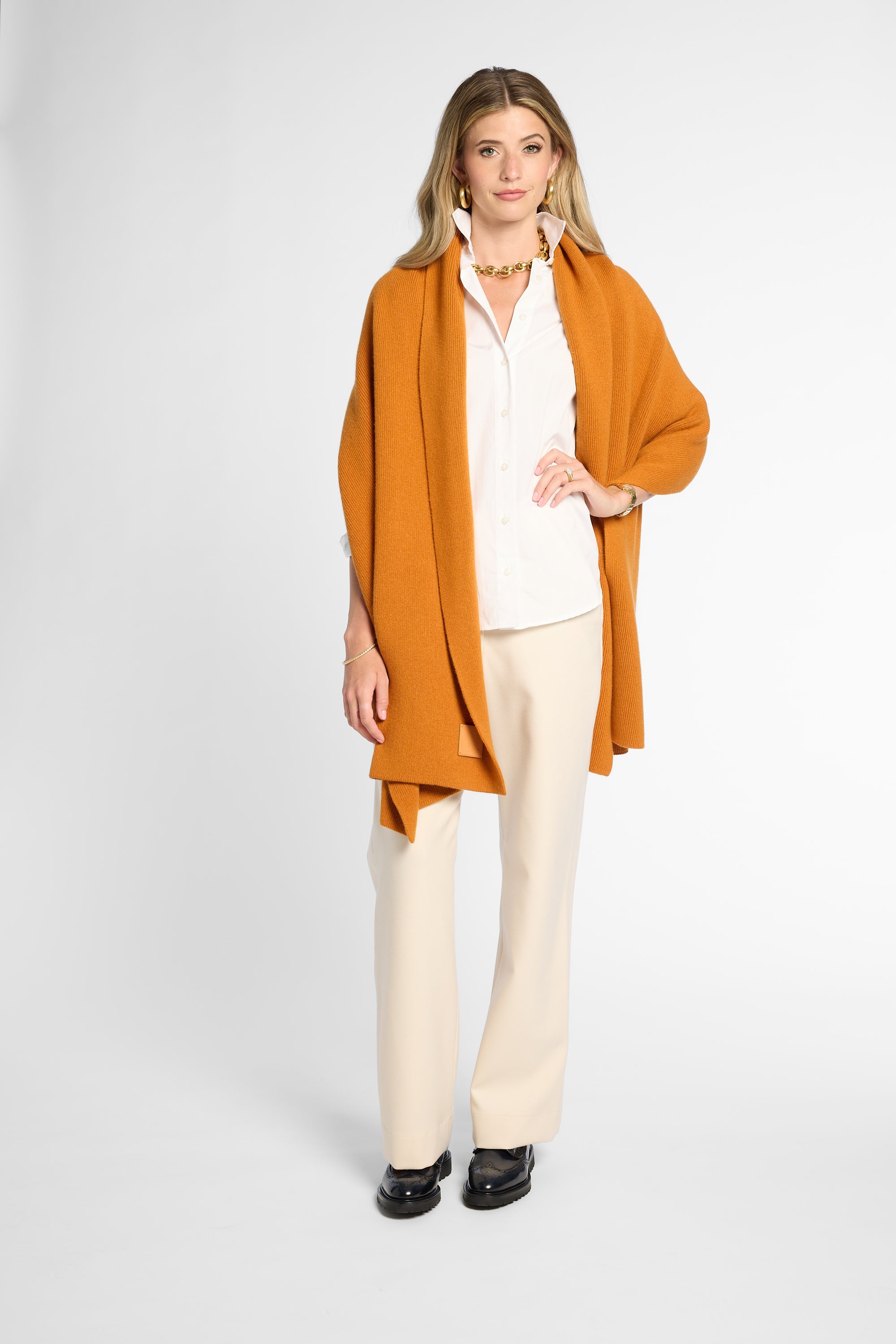A woman modeling a long-sleeved, open-front orange-colored wrap over a white collared shirt and cream wide-leg trousers, standing against a light gray studio background.
