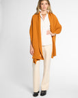 A woman modeling a long-sleeved, open-front orange-colored wrap over a white collared shirt and cream wide-leg trousers, standing against a light gray studio background.