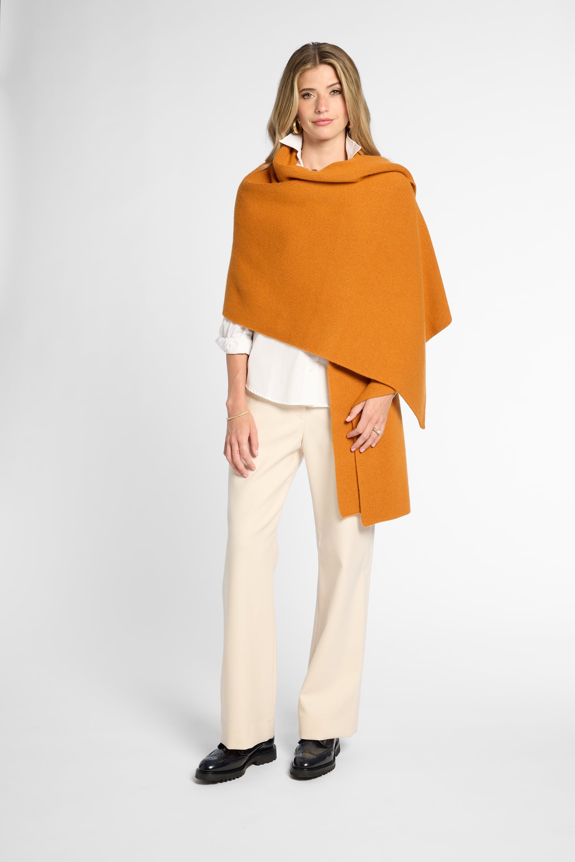 A woman modeling a draped orange orange wrap over a white blouse and cream trousers, standing against a light gray studio background.