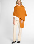 A woman modeling a draped orange orange wrap over a white blouse and cream trousers, standing against a light gray studio background.