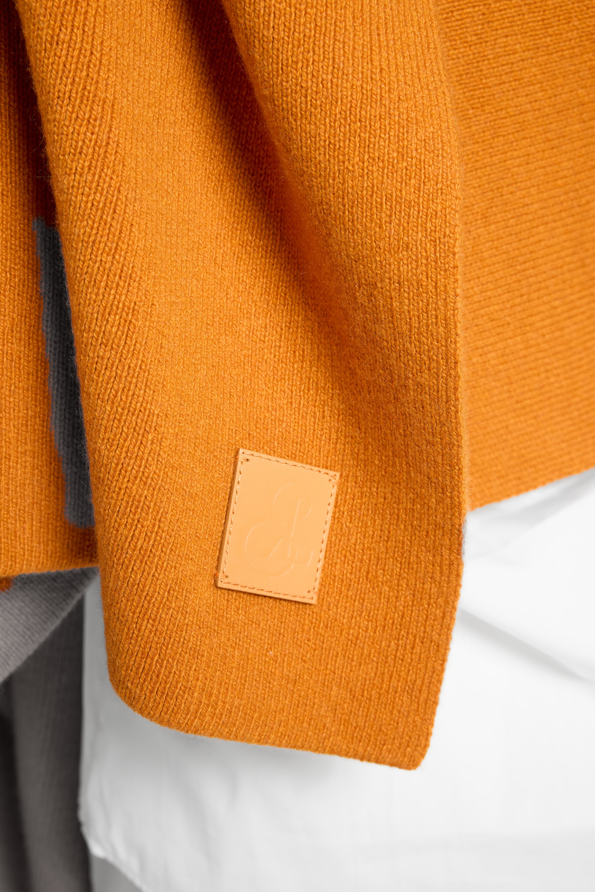 Close-up of a textured mustard orange knit wrap, with a small tan leather brand patch sewn onto the sleeve. The garment is styled over a white shirt and gray trousers.
