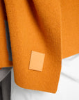 Close-up of a textured mustard orange knit wrap, with a small tan leather brand patch sewn onto the sleeve. The garment is styled over a white shirt and gray trousers.