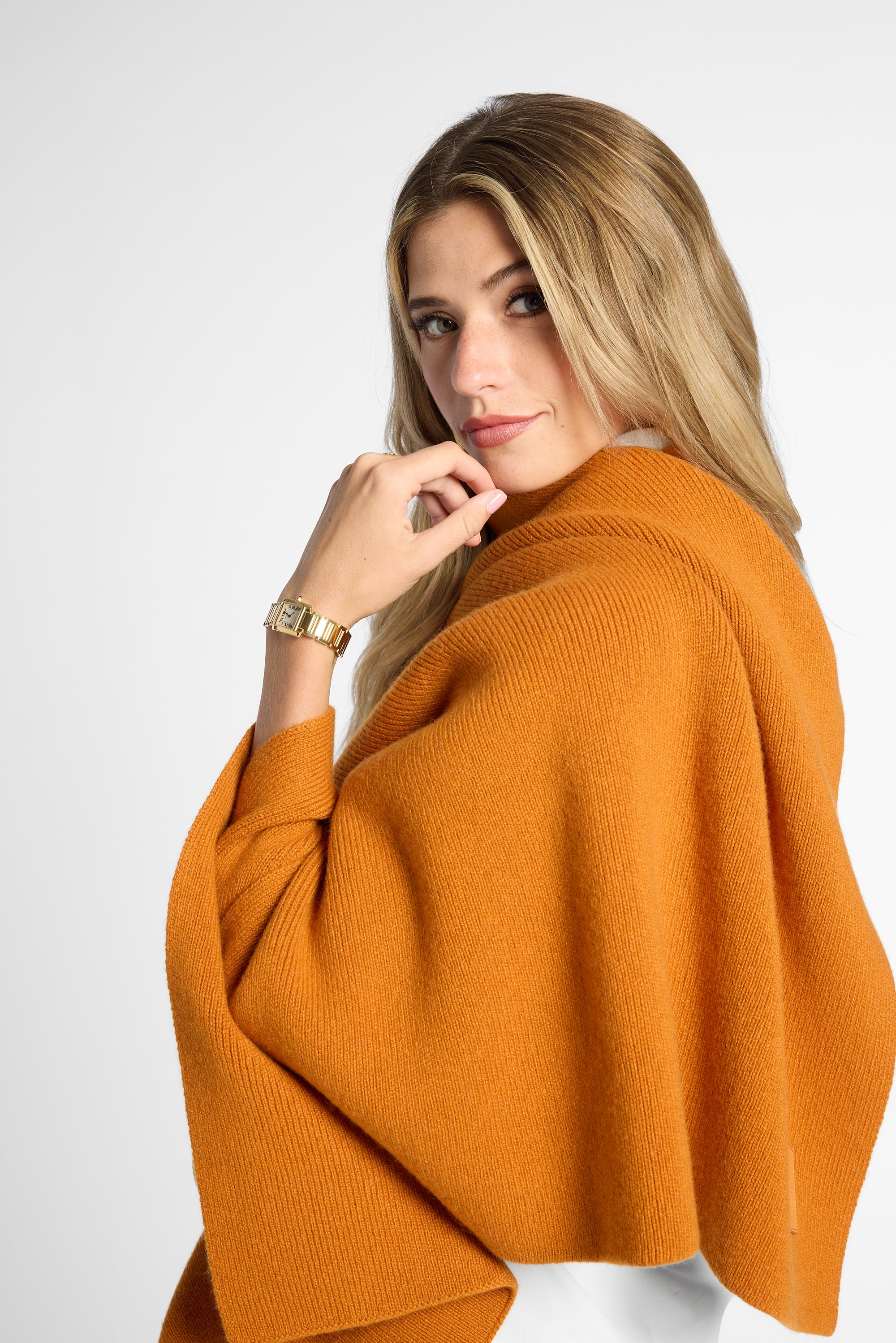 A woman with blonde hair wearing a warm orange knit poncho wrap, photographed against a clean light gray studio background.