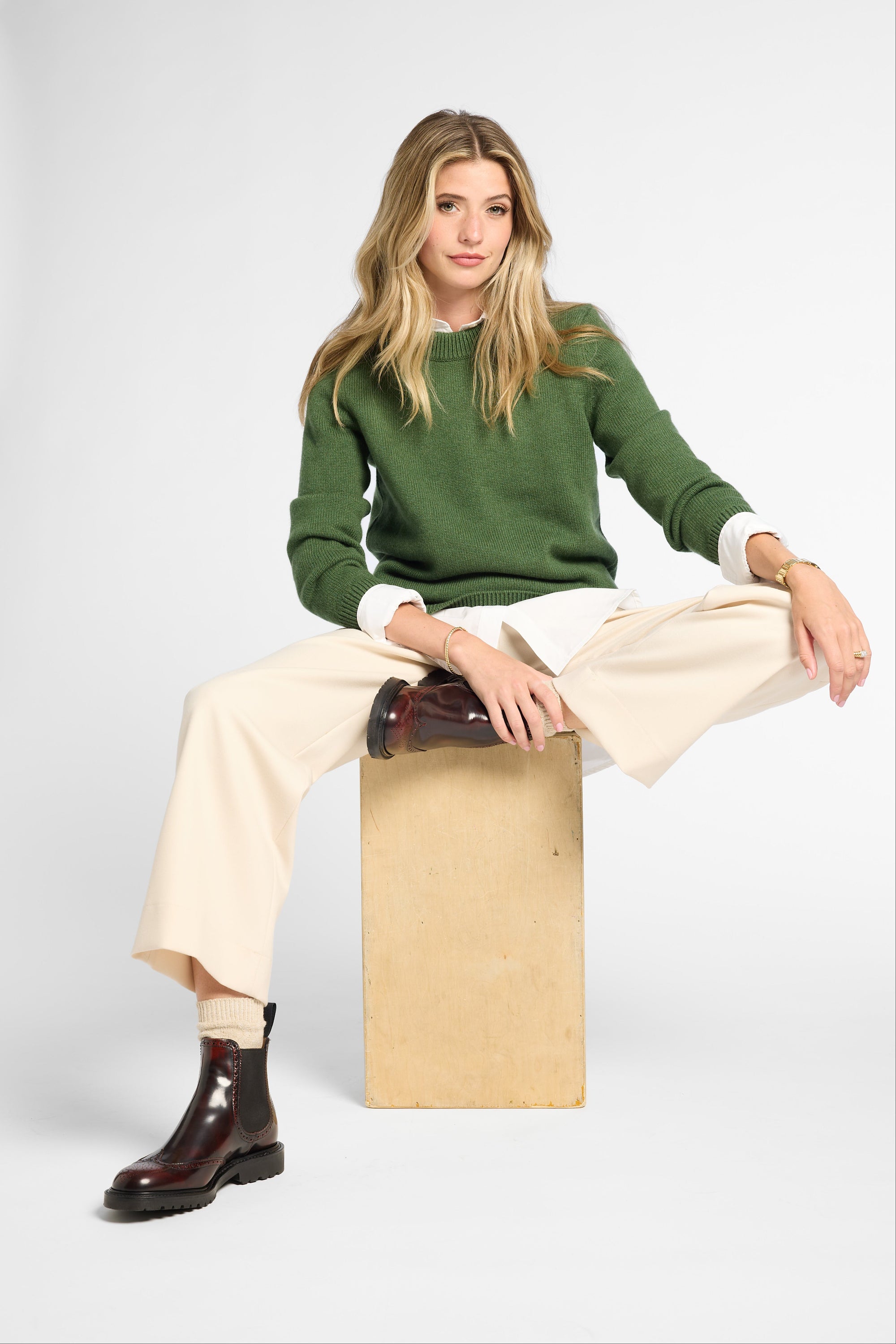 A woman models a green crewneck sweater, cream wide-leg trousers, and dark brown leather ankle boots while sitting on a light wood block against a white studio background.