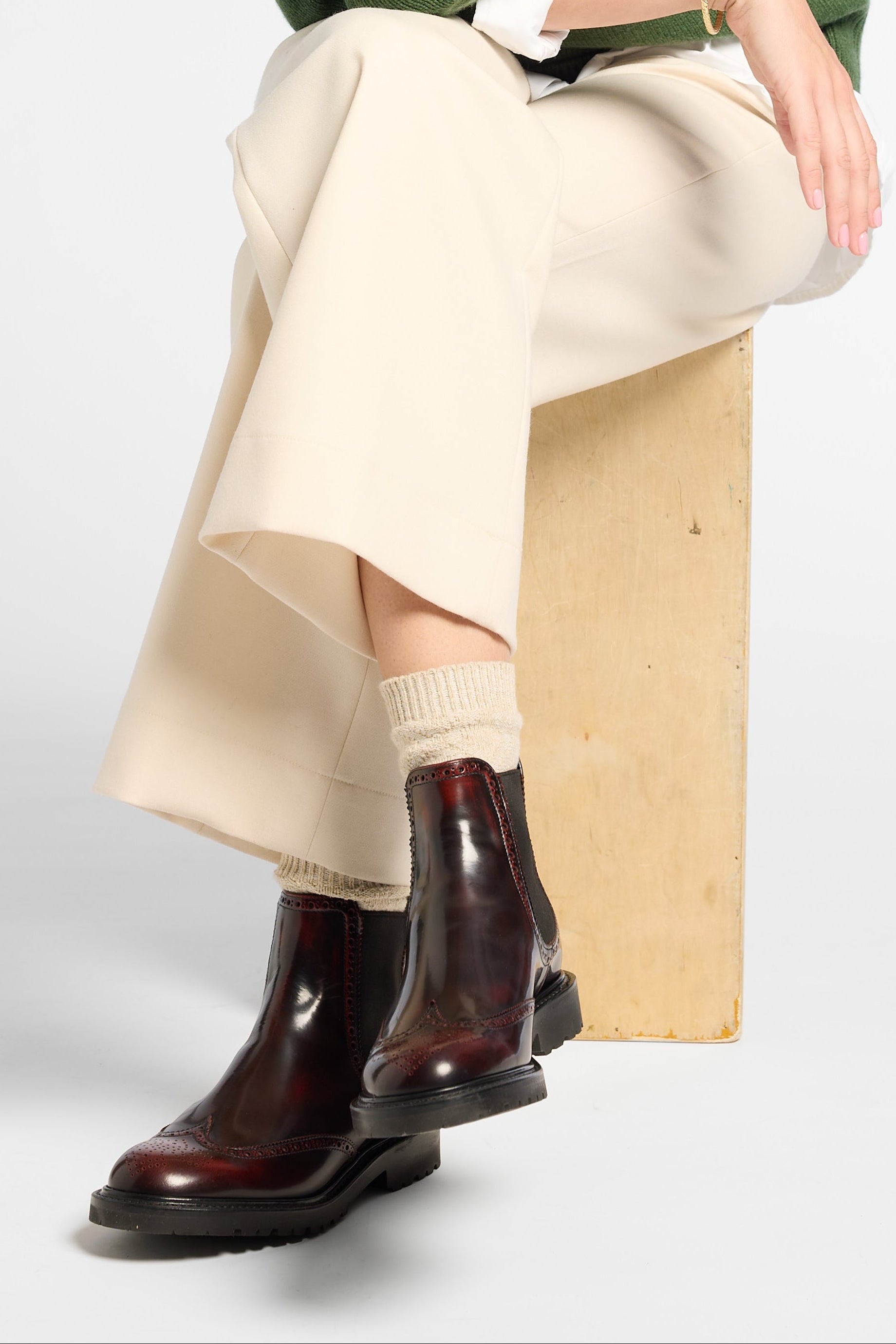 A woman sits on a wooden block wearing a green knit sweater over a white collared shirt, a cream pleated skirt, beige socks, and dark brown ankle boots, photographed against a light gray studio background.