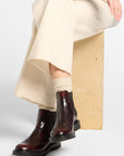 A woman sits on a wooden block wearing a green knit sweater over a white collared shirt, a cream pleated skirt, beige socks, and dark brown ankle boots, photographed against a light gray studio background.