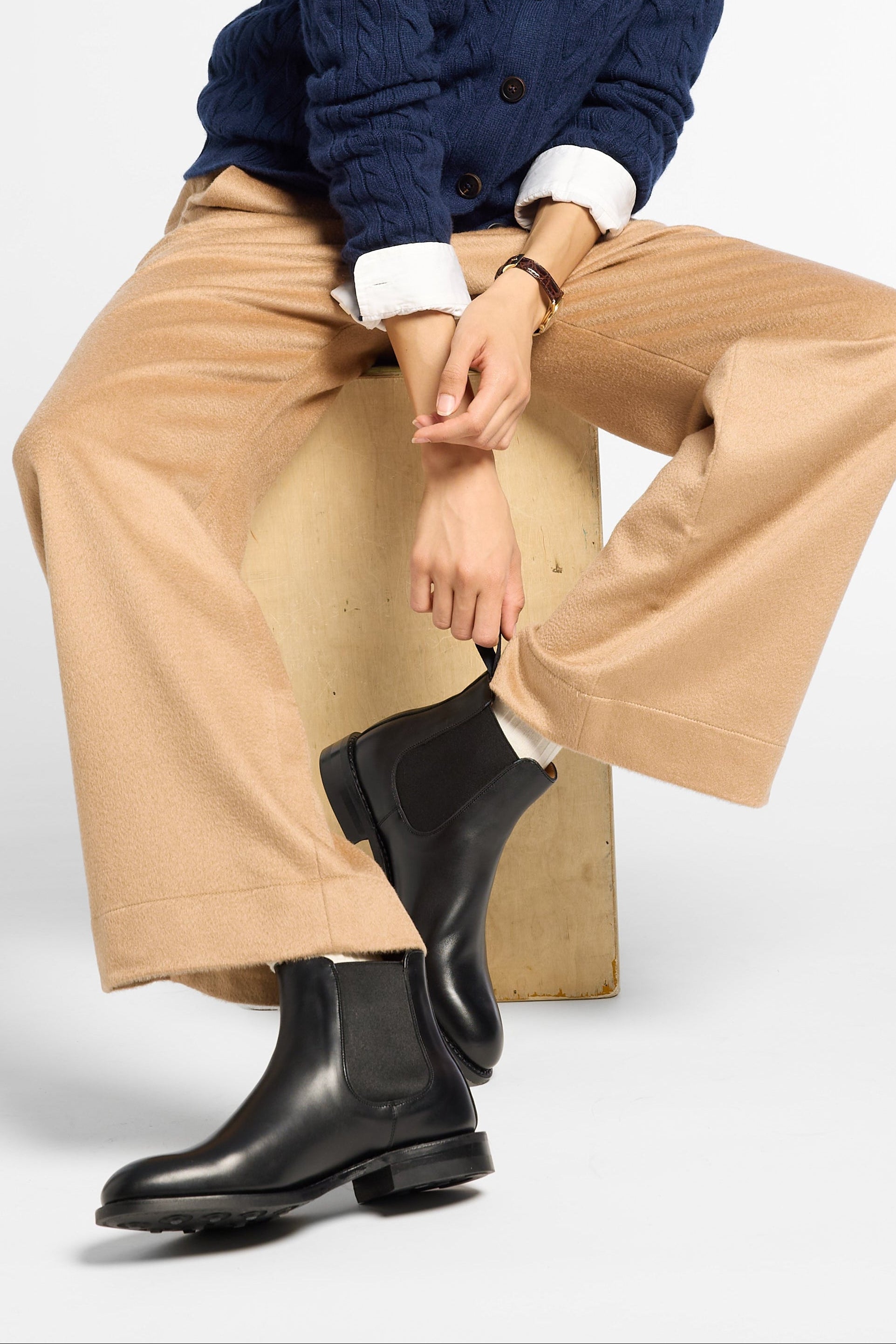 A model sits on a wooden block wearing a navy blue cable-knit cardigan over a white collared shirt, beige wide-leg trousers, and black Chelsea boots against a light gray studio background.