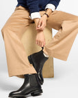 A model sits on a wooden block wearing a navy blue cable-knit cardigan over a white collared shirt, beige wide-leg trousers, and black Chelsea boots against a light gray studio background.